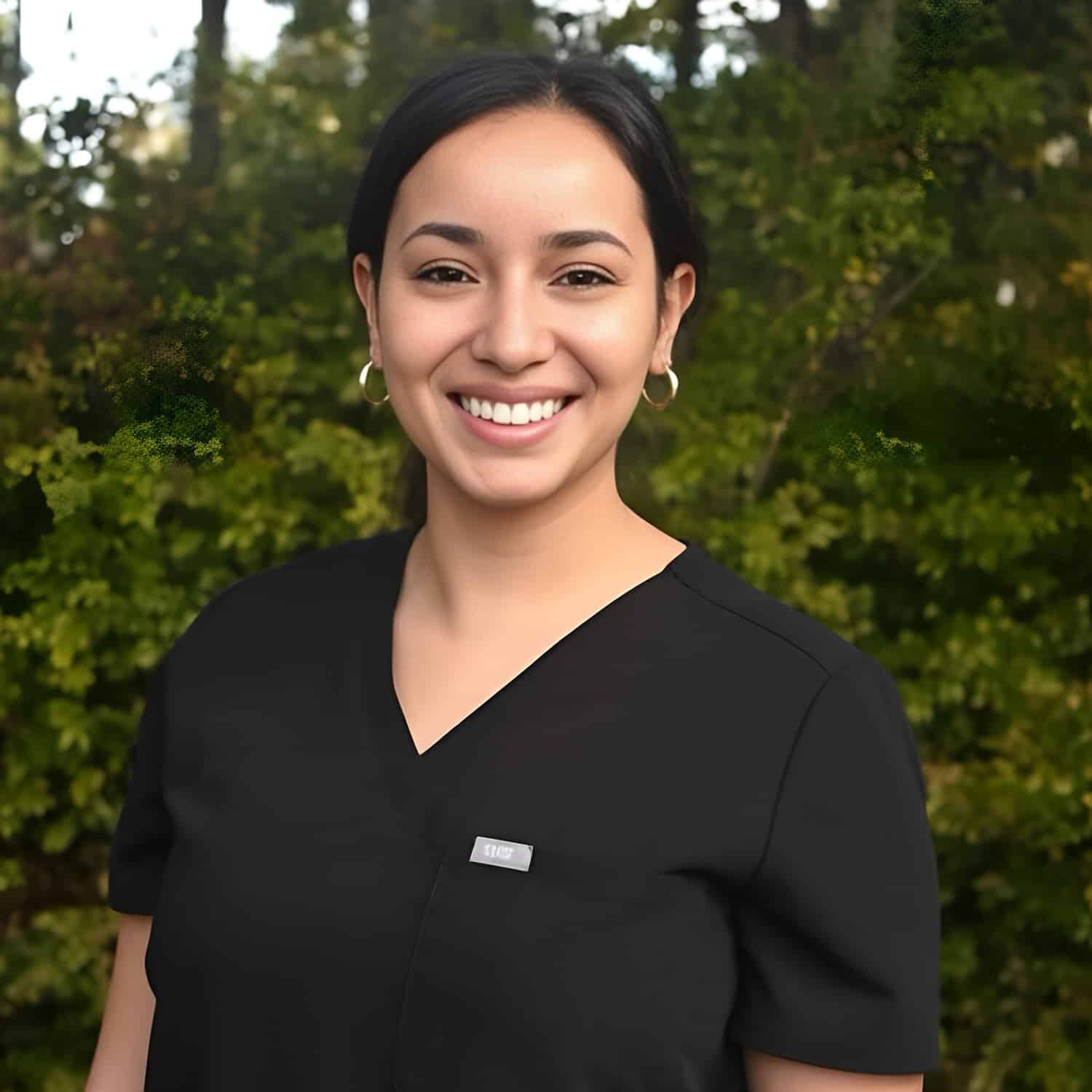 Smiling woman in black scrubs outdoors with greenery background.